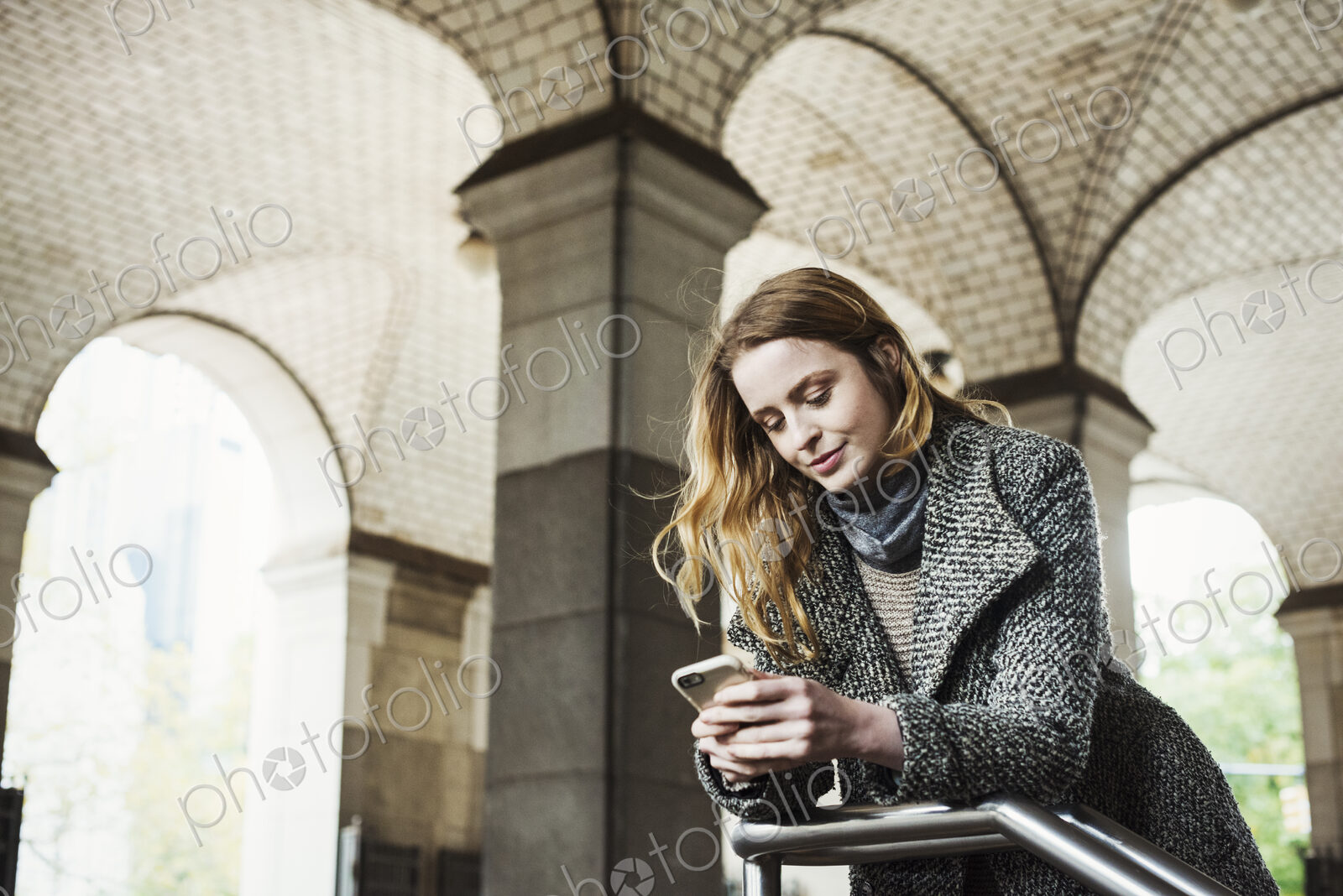 A woman with long hair looking at her smart phone, under an archway.