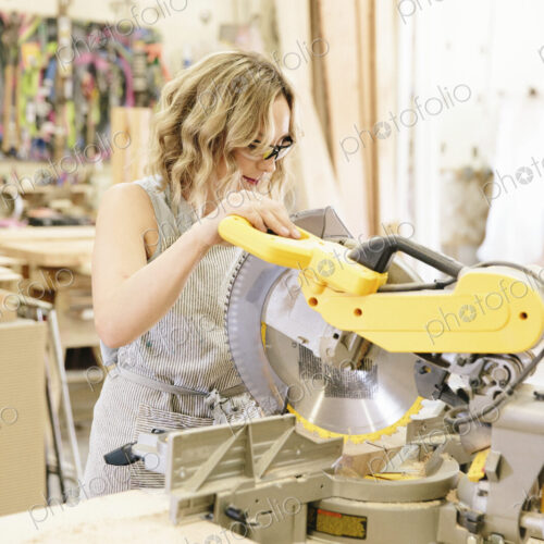 A woman woodworker using a mechanical saw to cut wood on a workbench.