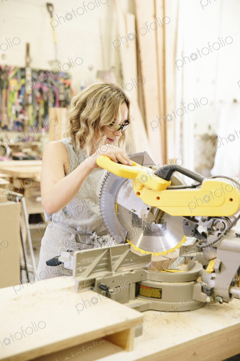 A woman woodworker using a mechanical saw to cut wood on a workbench.