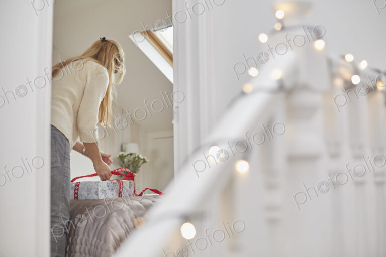 A woman wrapping red ribbons around Christmas presents on a bed, view through the…