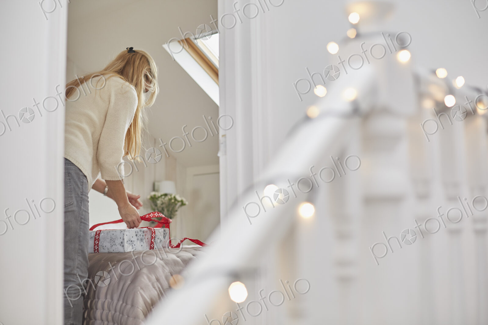 A woman wrapping red ribbons around Christmas presents on a bed, view through the…