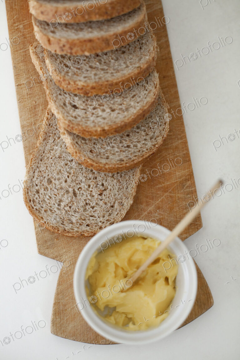 A wooden breadboard with a sliced brown loaf laid out. A dish of butter with a wooden…