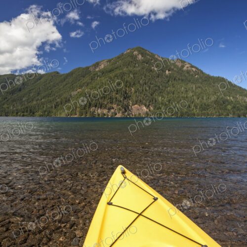 A yellow kayak rests on the clear shoreline of Lake Crescent with forested mountains…