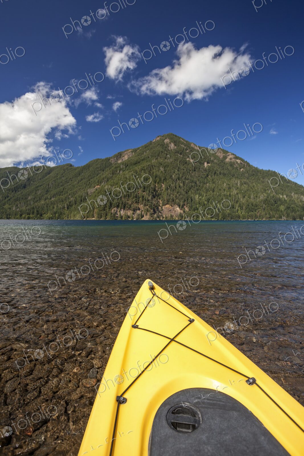 A yellow kayak rests on the clear shoreline of Lake Crescent with forested mountains…
