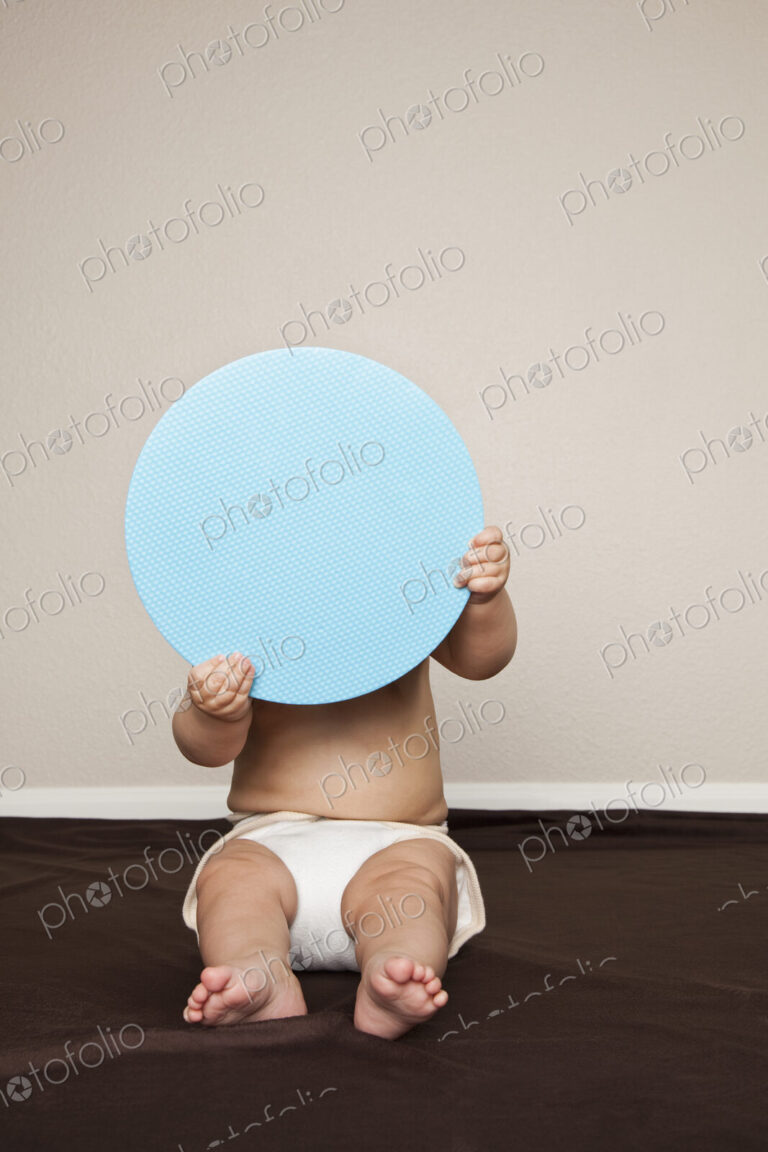 A young 8 month old baby boy wearing cloth diapers, hiding behind a large blue disc.