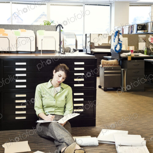 A young Caucasian businesswoman sitting on the floor of her office going over paperwork.