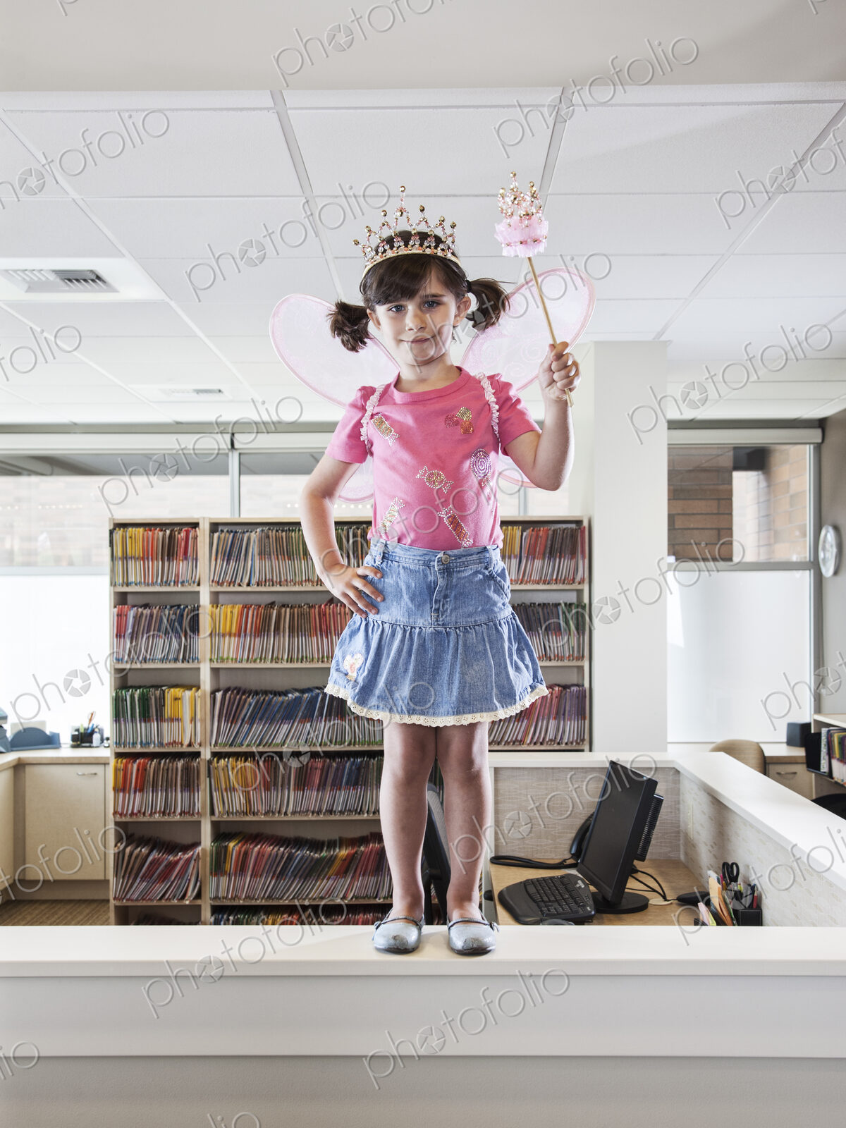 A young Caucasian girl dressed up as a fairy standing on a reception desk in a dental…