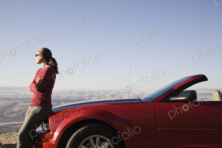 A young Caucasian woman leaning on her convertible sports car at a rest stop.