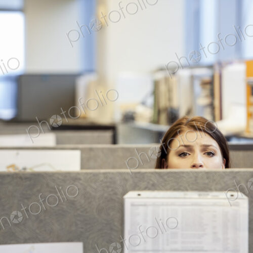 A young Caucasian woman looking concerned over the top of her cubicle in a corporate…