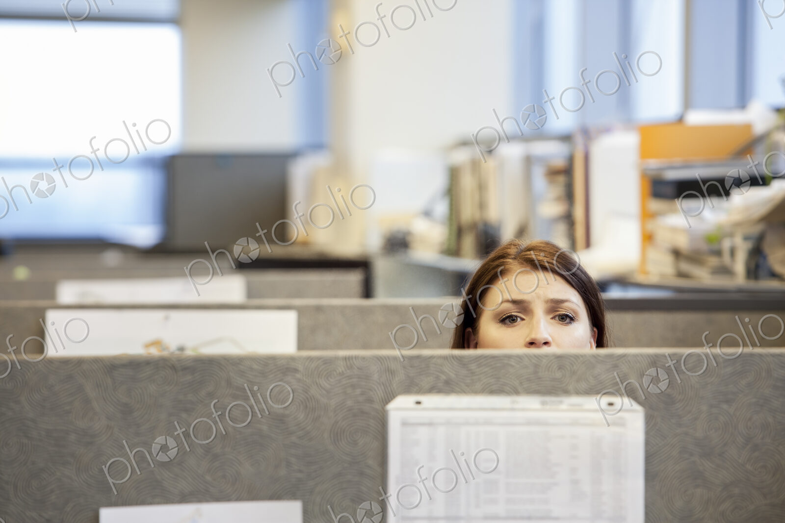 A young Caucasian woman looking concerned over the top of her cubicle in a corporate…