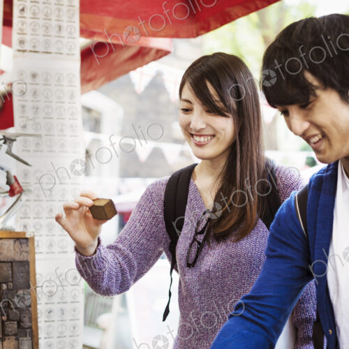 A young Japanese couple, man and woman at a souvenir street stall looking at printing…