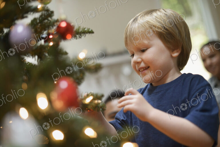 A young boy holding Christmas ornaments and placing them on the Christmas tree.