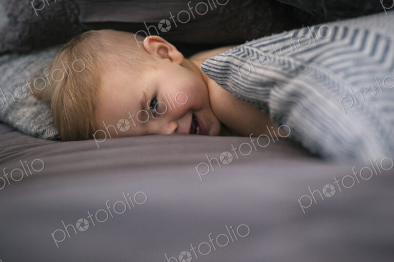 A young boy lying on his stomach in bed looking at the camera and laughing.