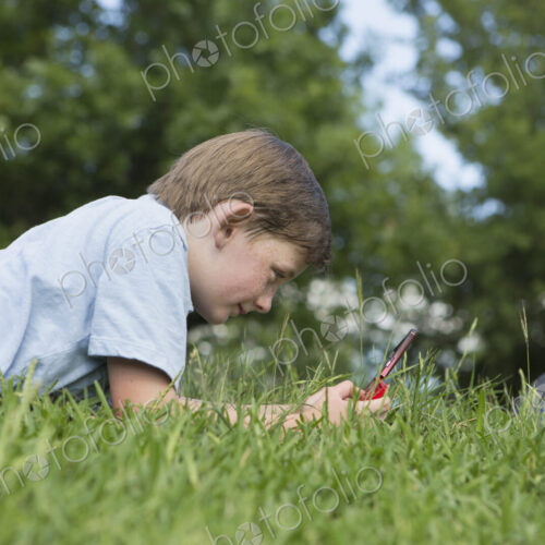 A young boy lying on the grass playing a hand held electronic game.