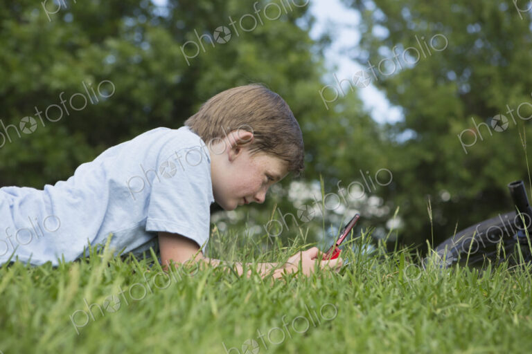 A young boy lying on the grass playing a hand held electronic game.