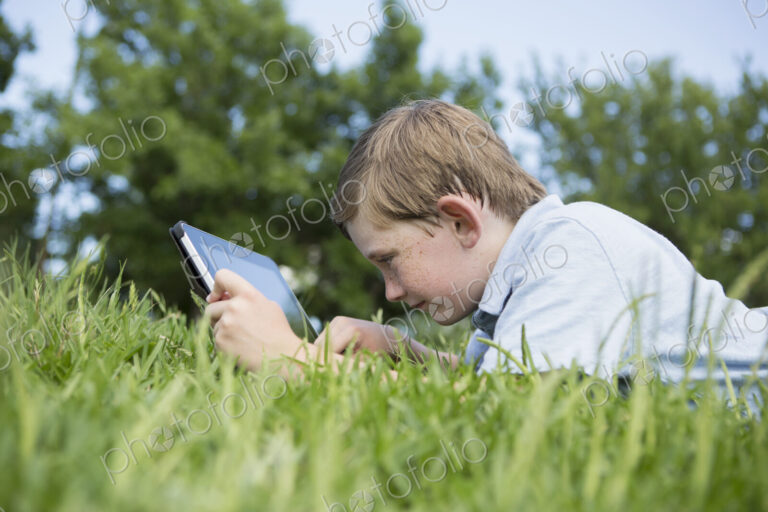A young boy lying on the grass, using a digital tablet.