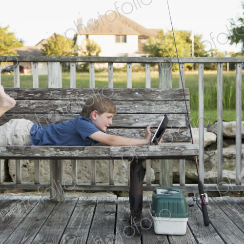 A young boy outdoors lying on a bench using a digital tablet. fishing equipment.