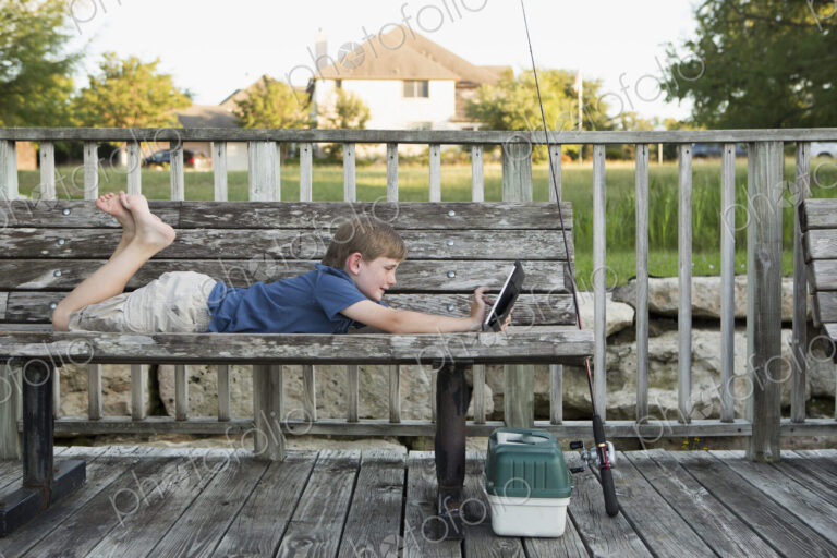 A young boy outdoors lying on a bench using a digital tablet. fishing equipment.