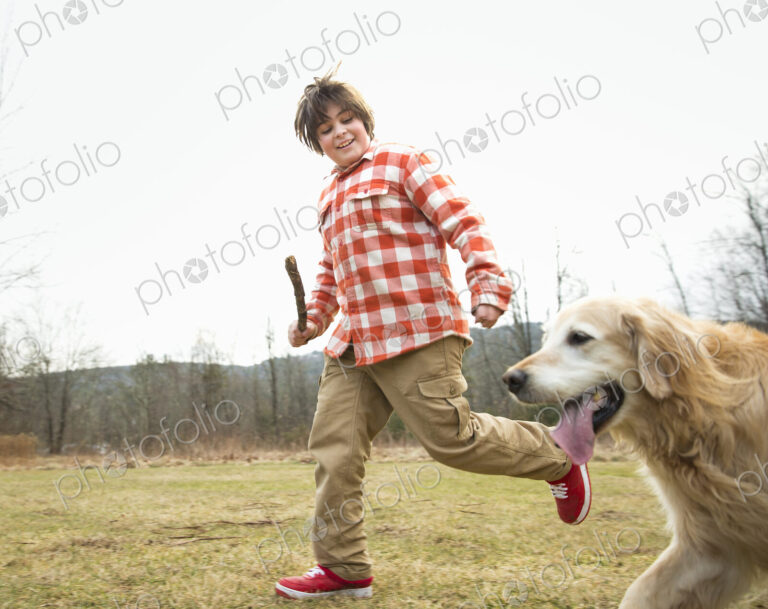 A young boy outdoors on a winter day, holding a stick and running with a golden…