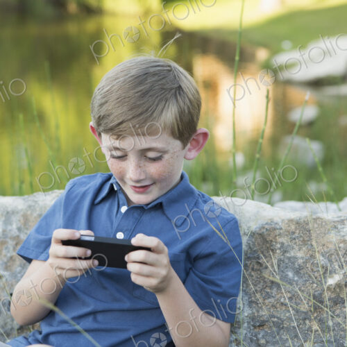 A young boy outdoors sitting leaning against a rock, using a handheld electronic game.