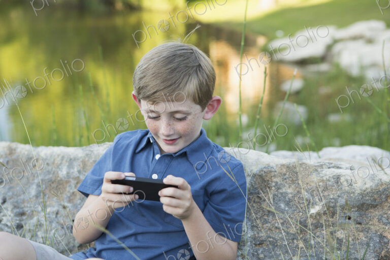 A young boy outdoors sitting leaning against a rock, using a handheld electronic game.