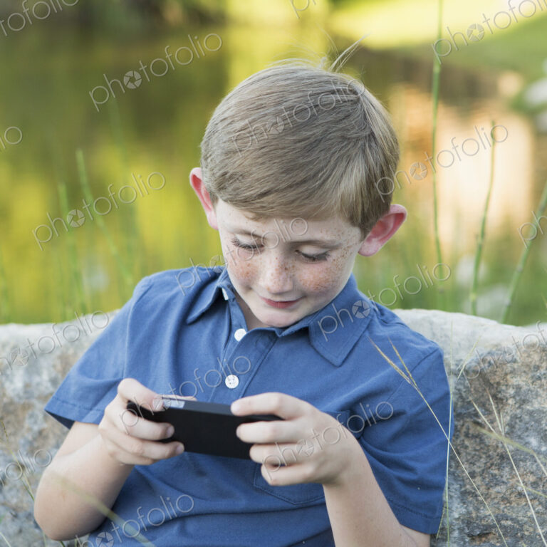 A young boy outdoors sitting leaning against a rock, using a handheld electronic game.