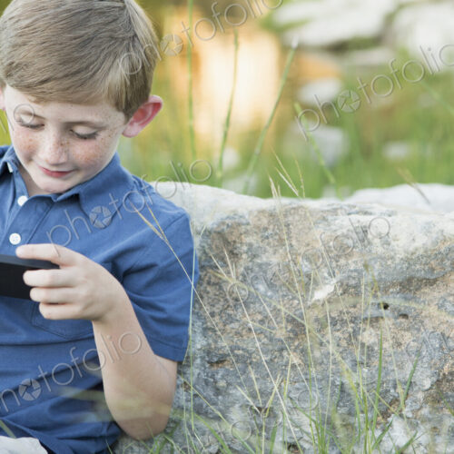 A young boy outdoors sitting leaning against a rock, using a handheld electronic game.