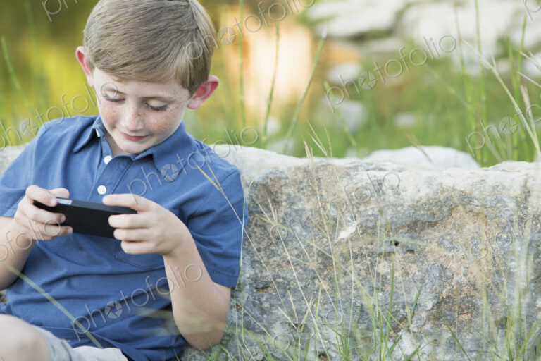 A young boy outdoors sitting leaning against a rock, using a handheld electronic game.