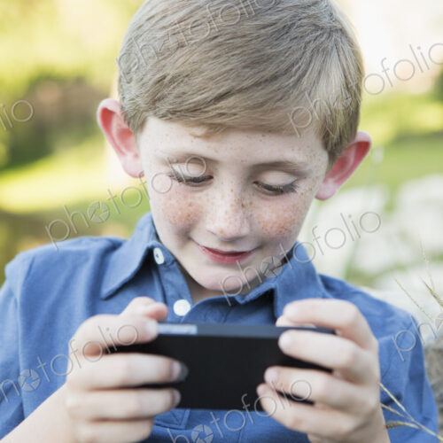 A young boy outdoors sitting leaning against a rock, using a handheld electronic game.
