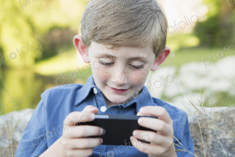 A young boy outdoors sitting leaning against a rock, using a handheld electronic game.