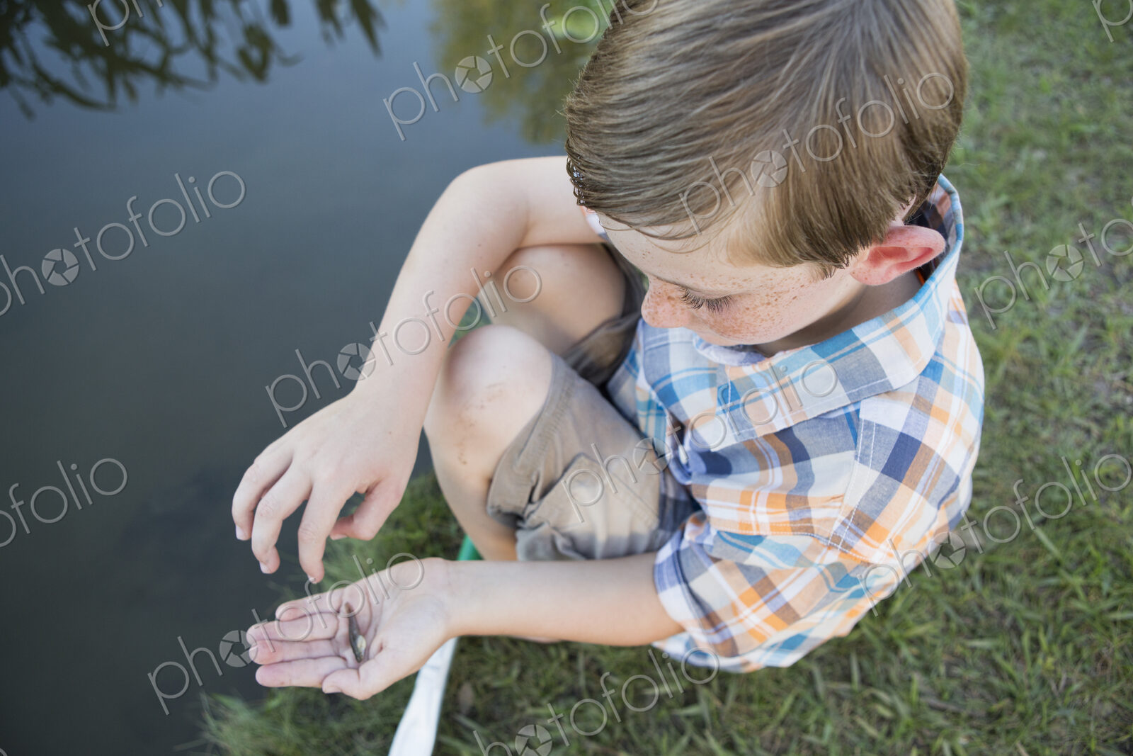 A young boy outdoors sitting on a riverbank with a small fish in the palm of his hand.