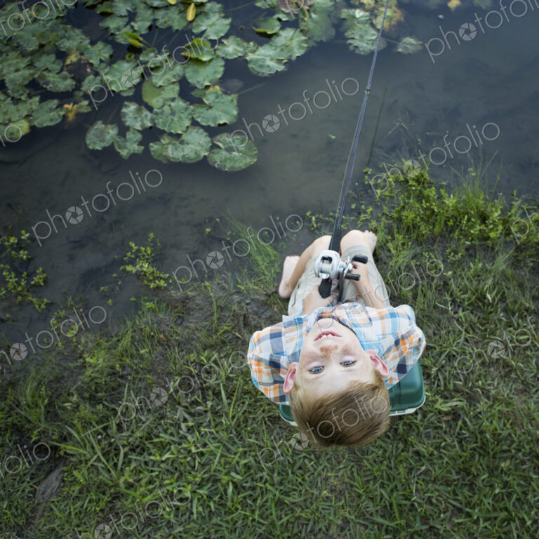 A young boy outdoors, standing on a riverbank looking up above his head.