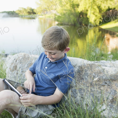 A young boy outdoors using a digital tablet. Sitting by water.