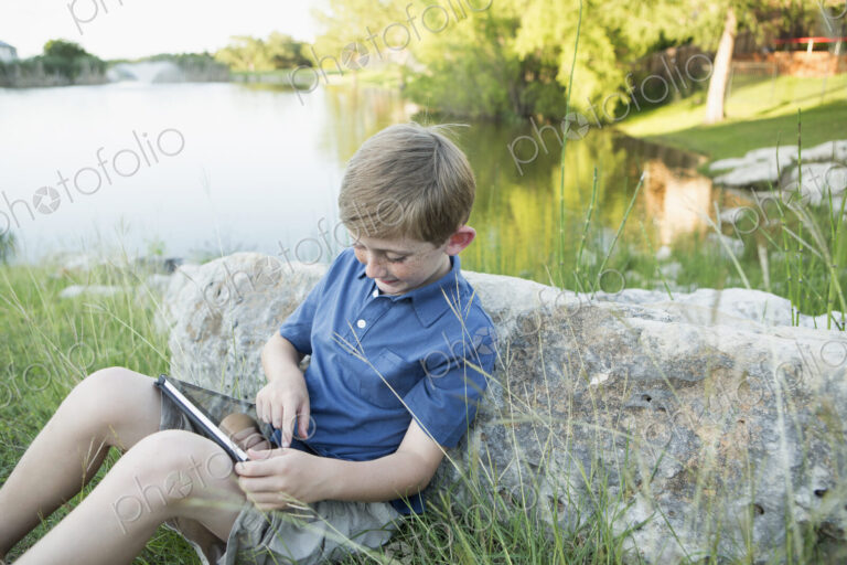 A young boy outdoors using a digital tablet. Sitting by water.
