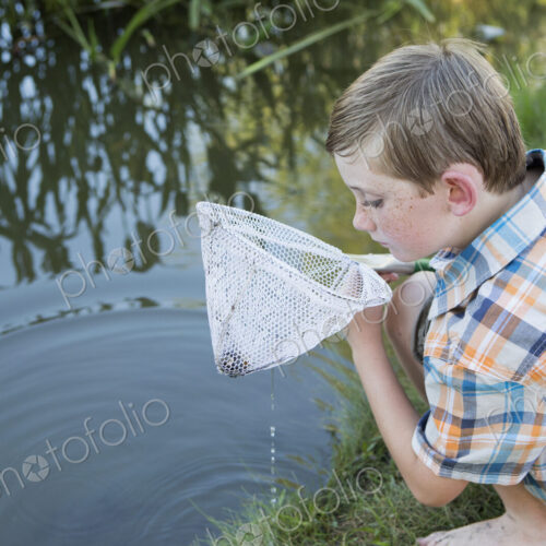 A young boy outdoors with a fishing net, examining the objects in the net, on a river…