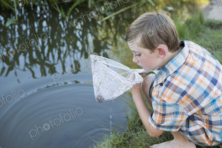 A young boy outdoors with a fishing net, examining the objects in the net, on a river…