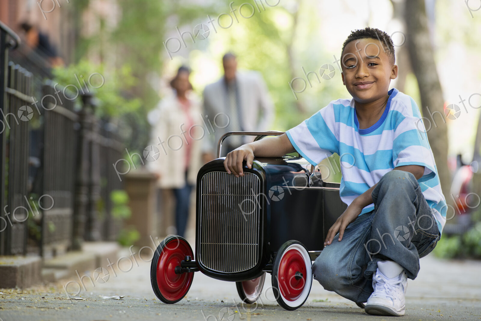 A young boy playing with a old fashioned toy car on wheels on a city street. A couple…
