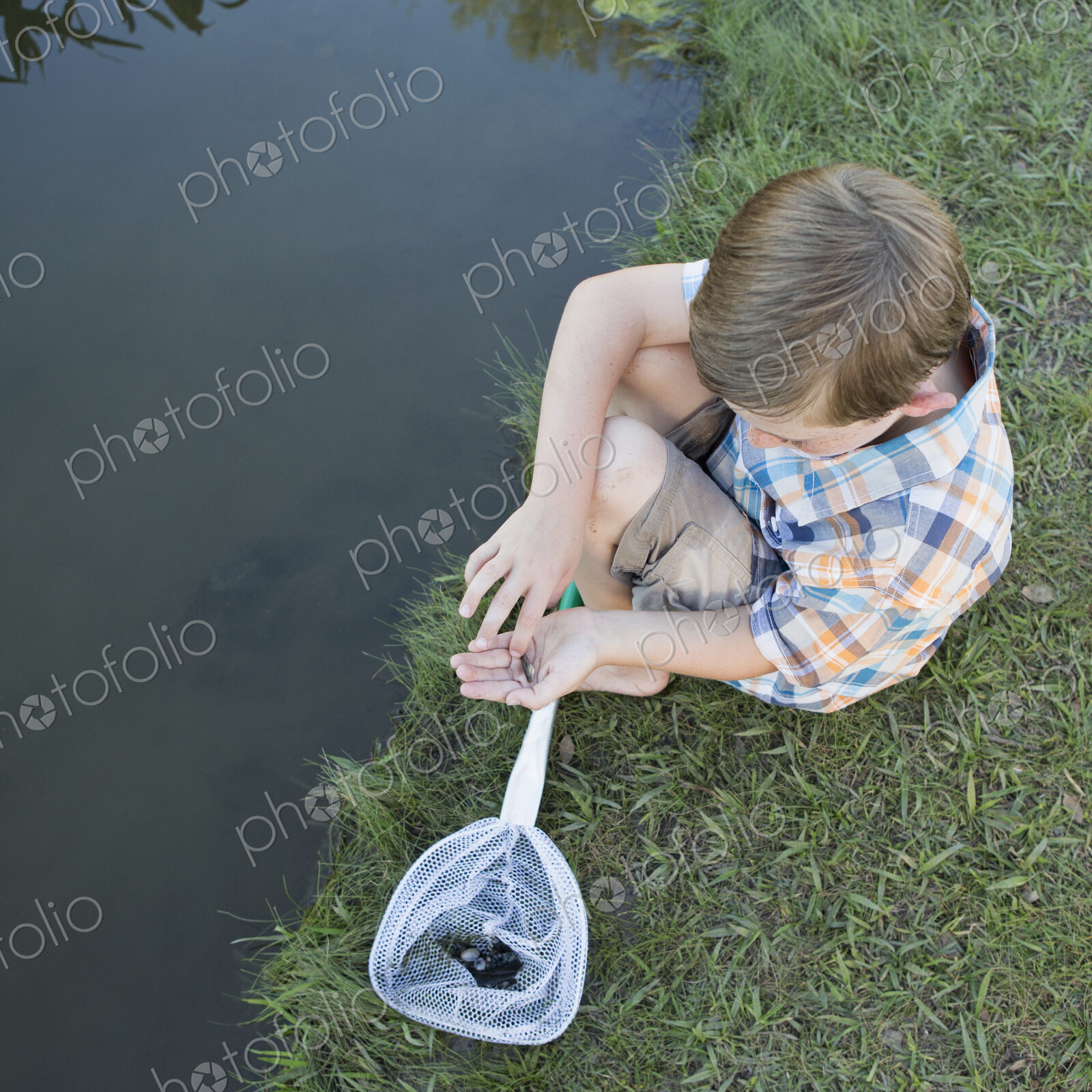 A young boy sitting on a river bank, examining a small fish, a tiddler he scooped up…