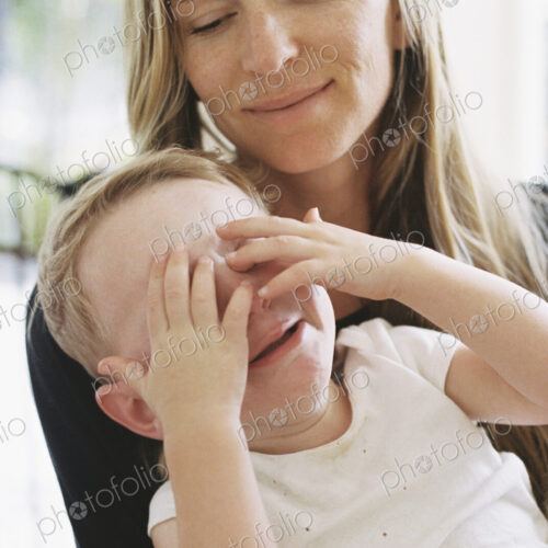 A young boy sitting on his mothers lap with his hands over his eyes.