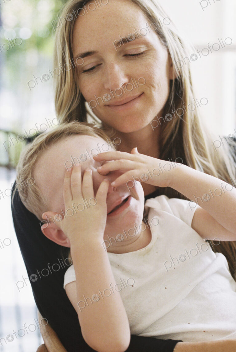A young boy sitting on his mothers lap with his hands over his eyes.