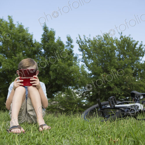 A young boy sitting on the grass playing a hand held electronic game.