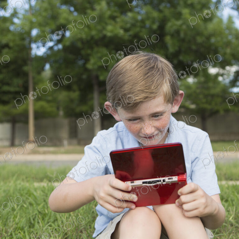 A young boy sitting on the grass playing a hand held electronic game.