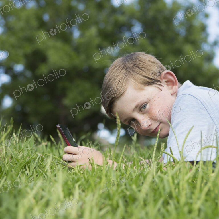 A young boy sitting on the grass using a hand held electronic games device.