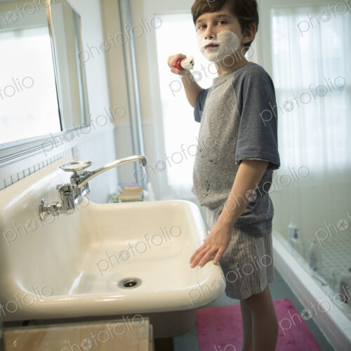 A young boy standing in a bathroom holding a shaving brush, covering his face with foam. A young boy standing in a bathroom holding a shaving brush, covering his face with foam.