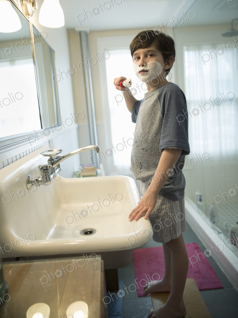A young boy standing in a bathroom holding a shaving brush, covering his face with foam.