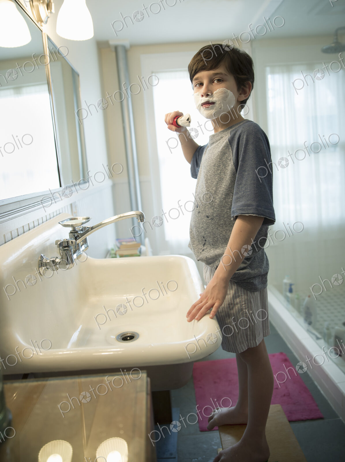 A young boy standing in a bathroom holding a shaving brush, covering his face with foam.