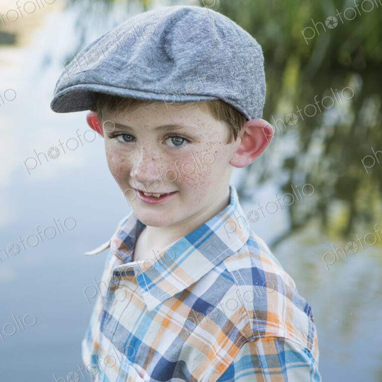 A young boy wearing a checked shirt and cloth cap with a large brim standing on a…