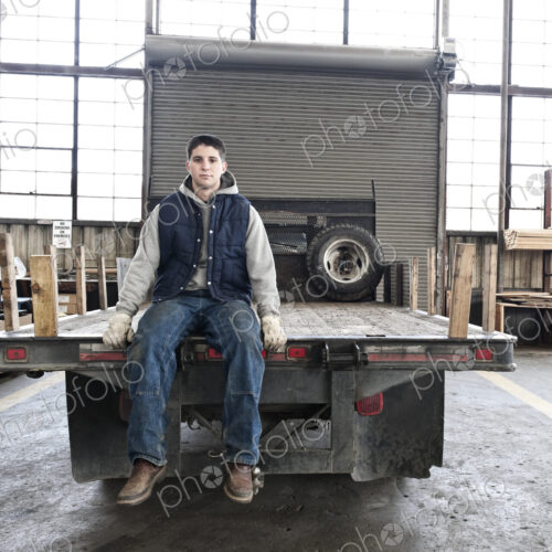 A young caucasian factory worker sitting on the back of a flat bed truck in a sheet…