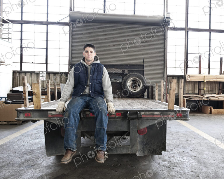 A young caucasian factory worker sitting on the back of a flat bed truck in a sheet…
