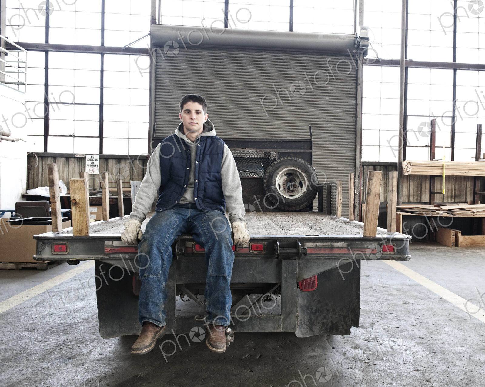 A young caucasian factory worker sitting on the back of a flat bed truck in a sheet…
